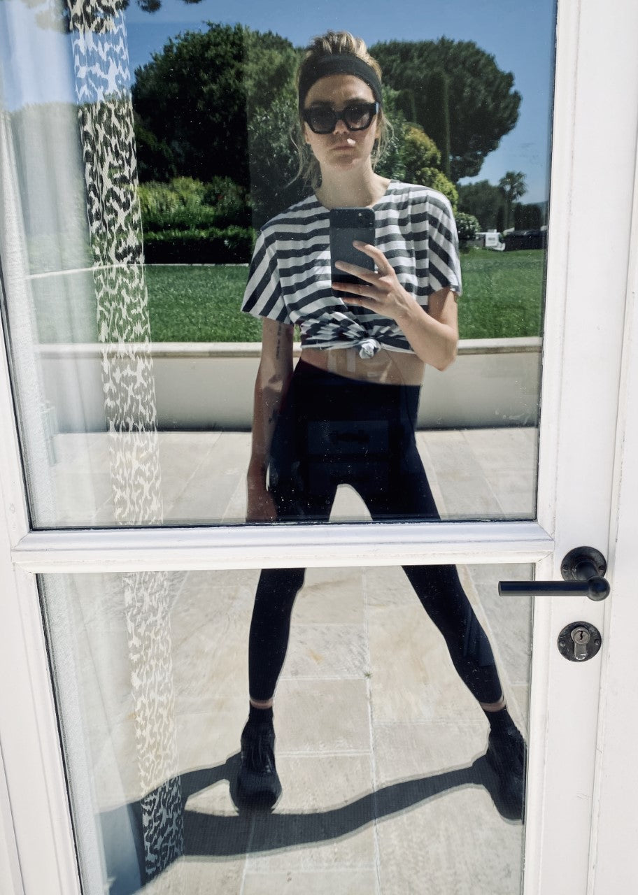 Person wearing sunglasses, striped shirt, and leggings takes a selfie in a glass door reflection with outdoor greenery in the background.