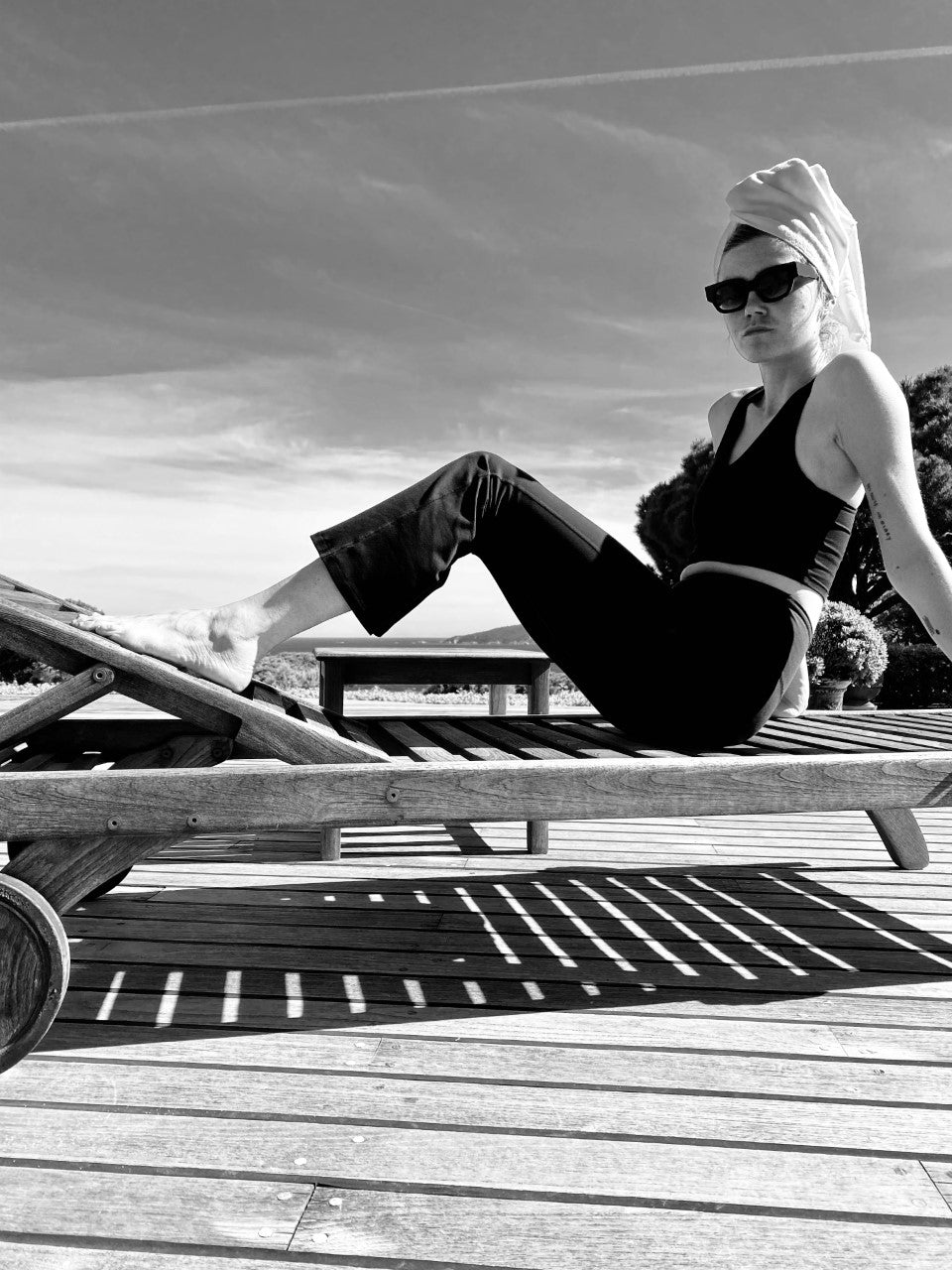 A person wearing sunglasses and a towel on their head lounges on a wooden chair outdoors under a clear sky.