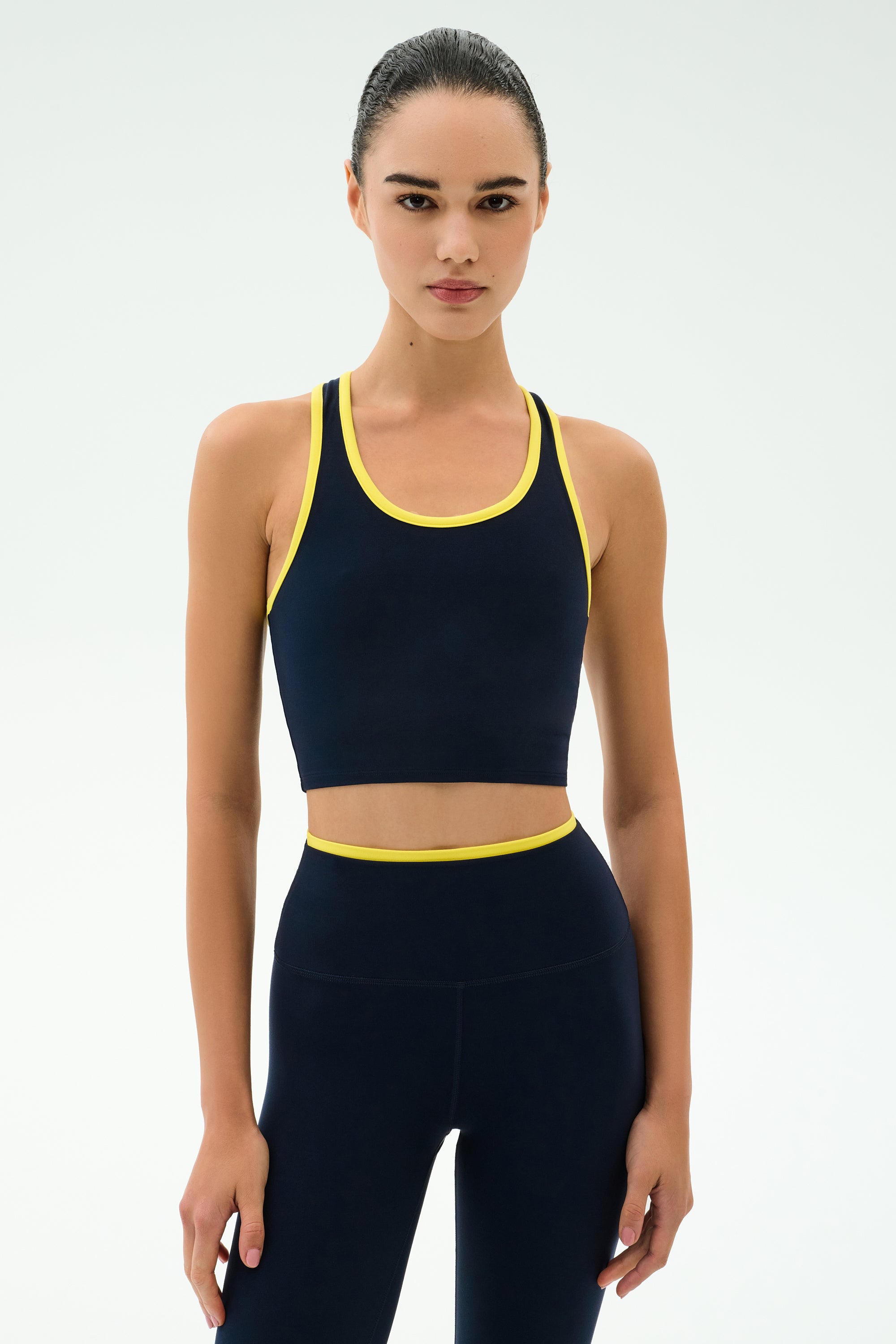 A woman stands against a plain background wearing the SPLITS59 Amber Airweight Bralette in Indigo/Ray, featuring a cropped scoop-neck style in performance fabric with yellow trim, looking directly at the camera.