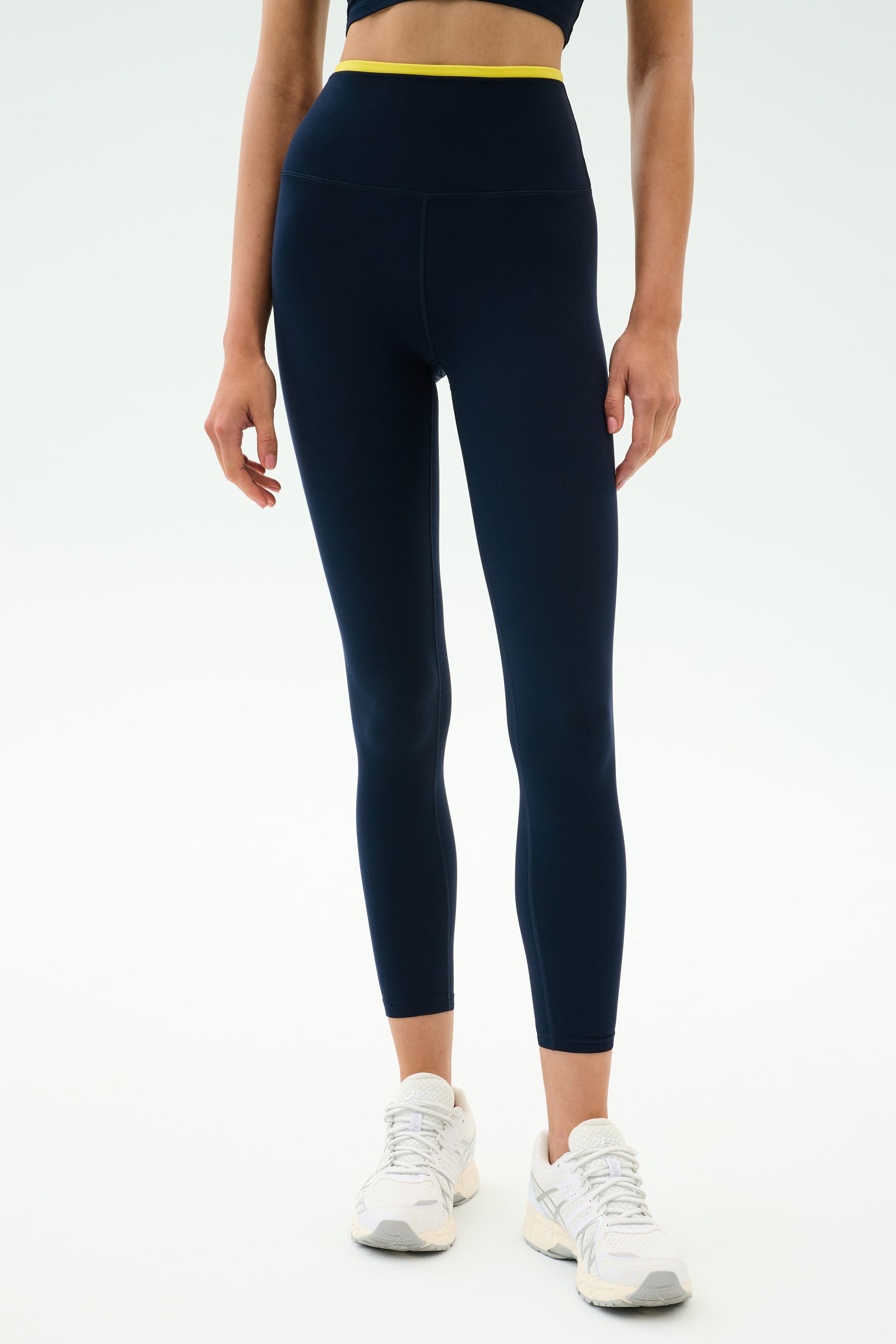A person stands against a white background in SPLITS59 Dual High Waist Airweight 7/8 leggings in Indigo/Ray—navy with a yellow waistband—paired with white sneakers and a dark crop top, ideal for hot yoga sessions.