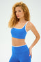 A person with curly hair poses confidently in the SPLITS59 Cait Rigor Bra in Classic Blue/White, paired with a matching high-performance outfit, against a plain background.