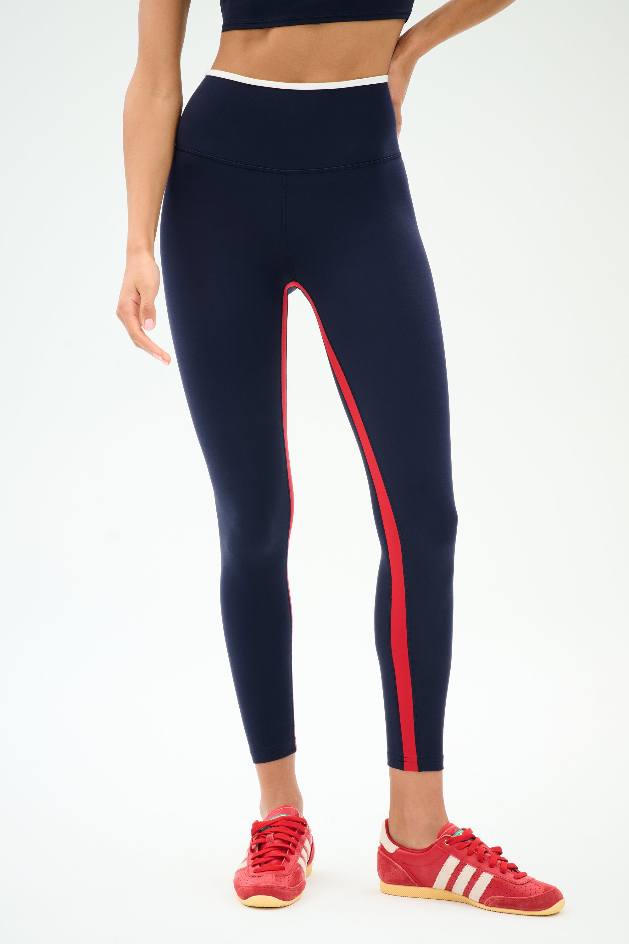 Model wears SPLITS59 Easton Rigor High Waist 7/8 leggings in Indigo/White, paired with a black sports bra and red Adidas sneakers. These leggings feature compressive fabric for enhanced workout support against a plain background.