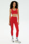 Woman against a white background, wearing the SPLITS59 Ella Airweight Bra in Pirate Red/Indigo, matching red leggings with side stripes, white socks, and gray sneakers.