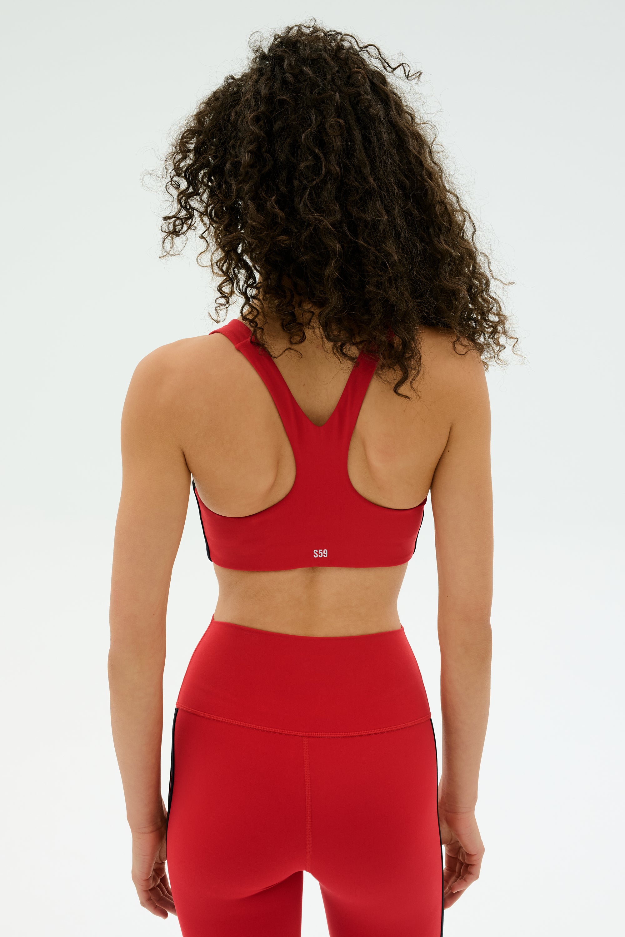 A person with curly hair is shown from the back, wearing the SPLITS59 Ella Airweight Bra in Pirate Red/Indigo against a plain light background.