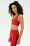 A person with curly hair wears the SPLITS59 Ella Airweight Bra in Pirate Red/Indigo and matching leggings, featuring black side stripes, standing against a plain white background.