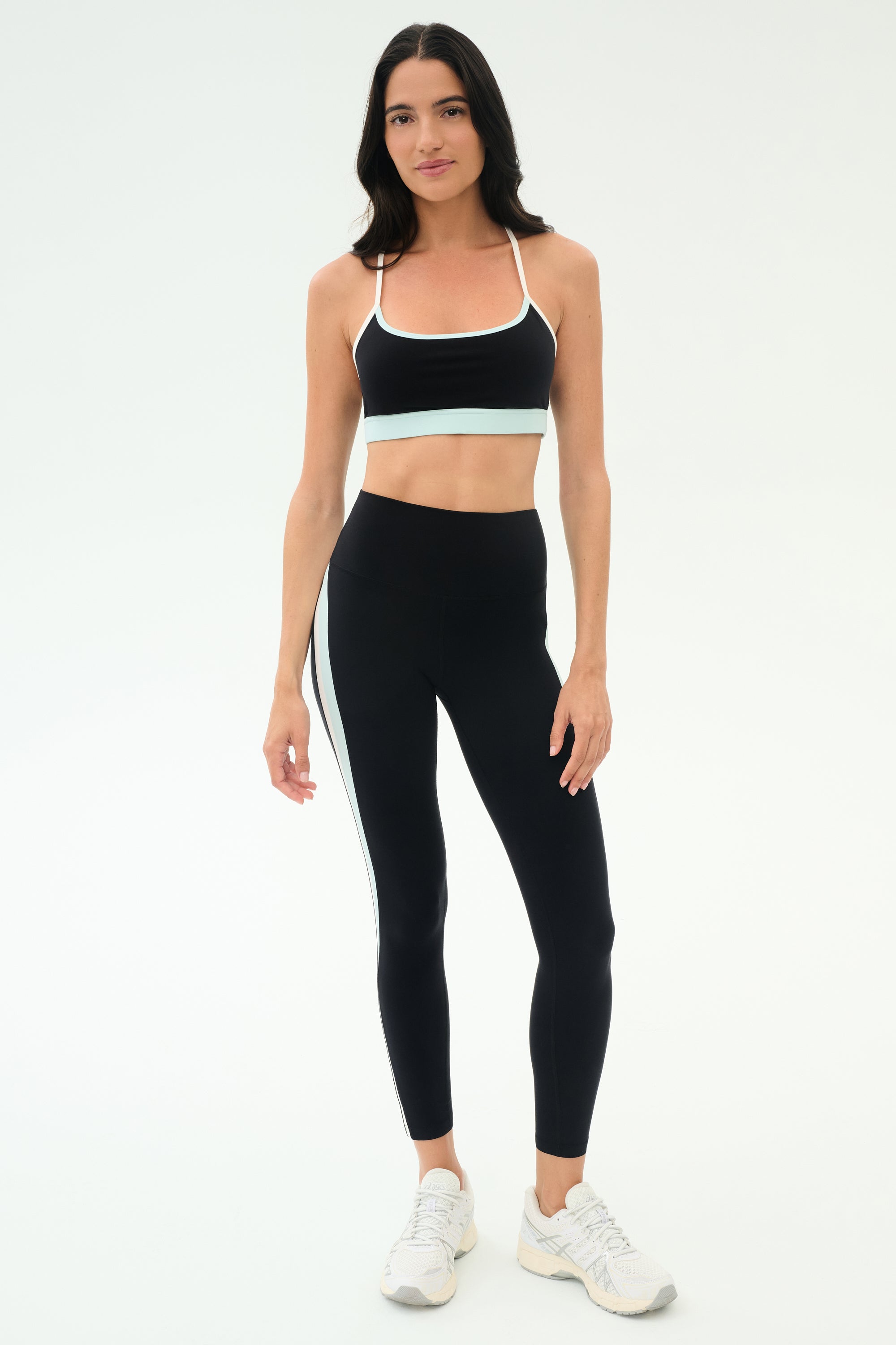 A woman faces forward in a black sports bra, SPLITS59 Emerson Airweight High Waist leggings in Black/Ice with a white side stripe—ideal for hot yoga or Pilates—and white athletic shoes.