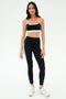 A woman faces forward in a black sports bra, SPLITS59 Emerson Airweight High Waist leggings in Black/Ice with a white side stripe—ideal for hot yoga or Pilates—and white athletic shoes.