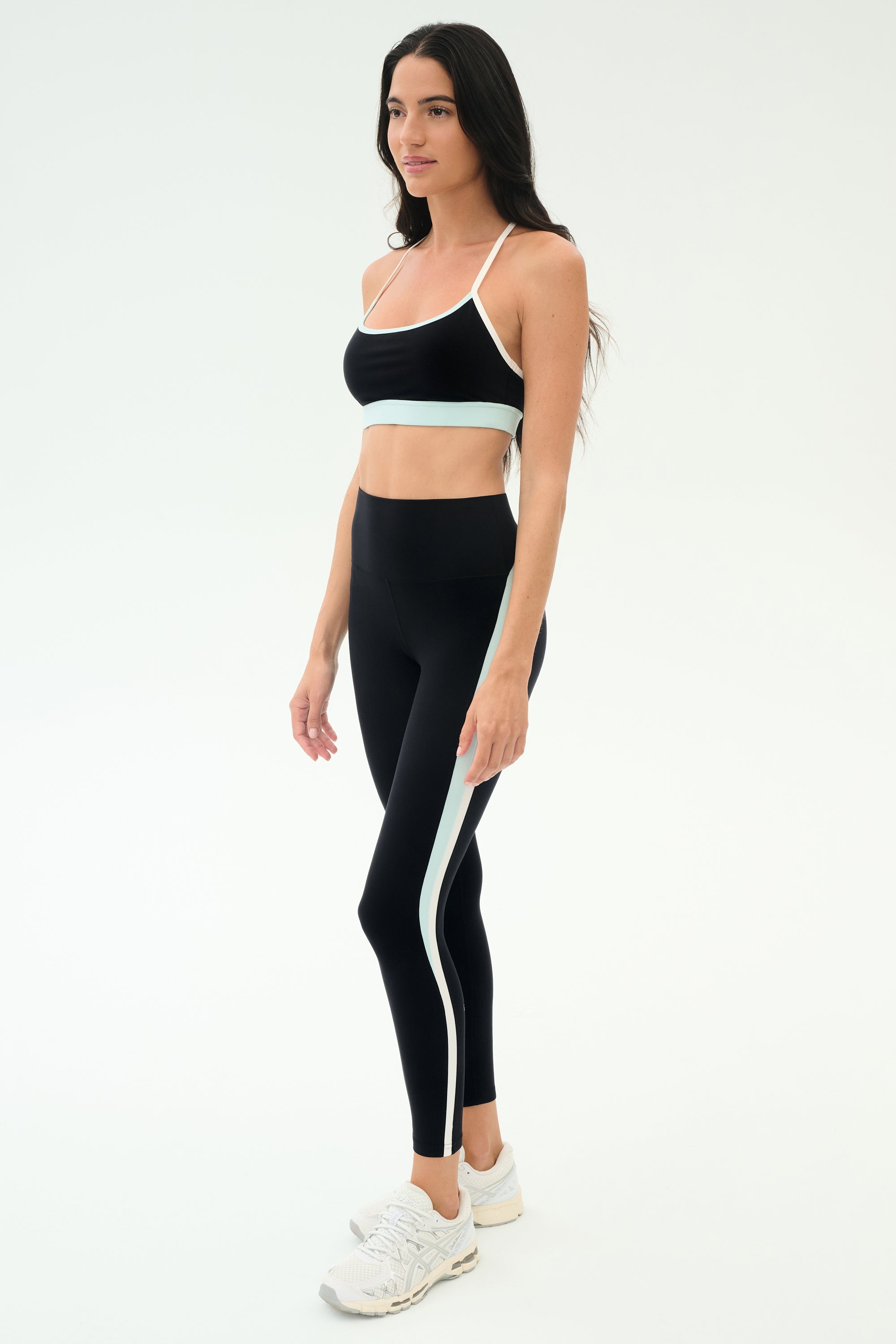 A woman wears the SPLITS59 Emerson Airweight High Waist leggings in Black/Ice and a black sports bra, ideal for Pilates or hot yoga, paired with white athletic shoes.