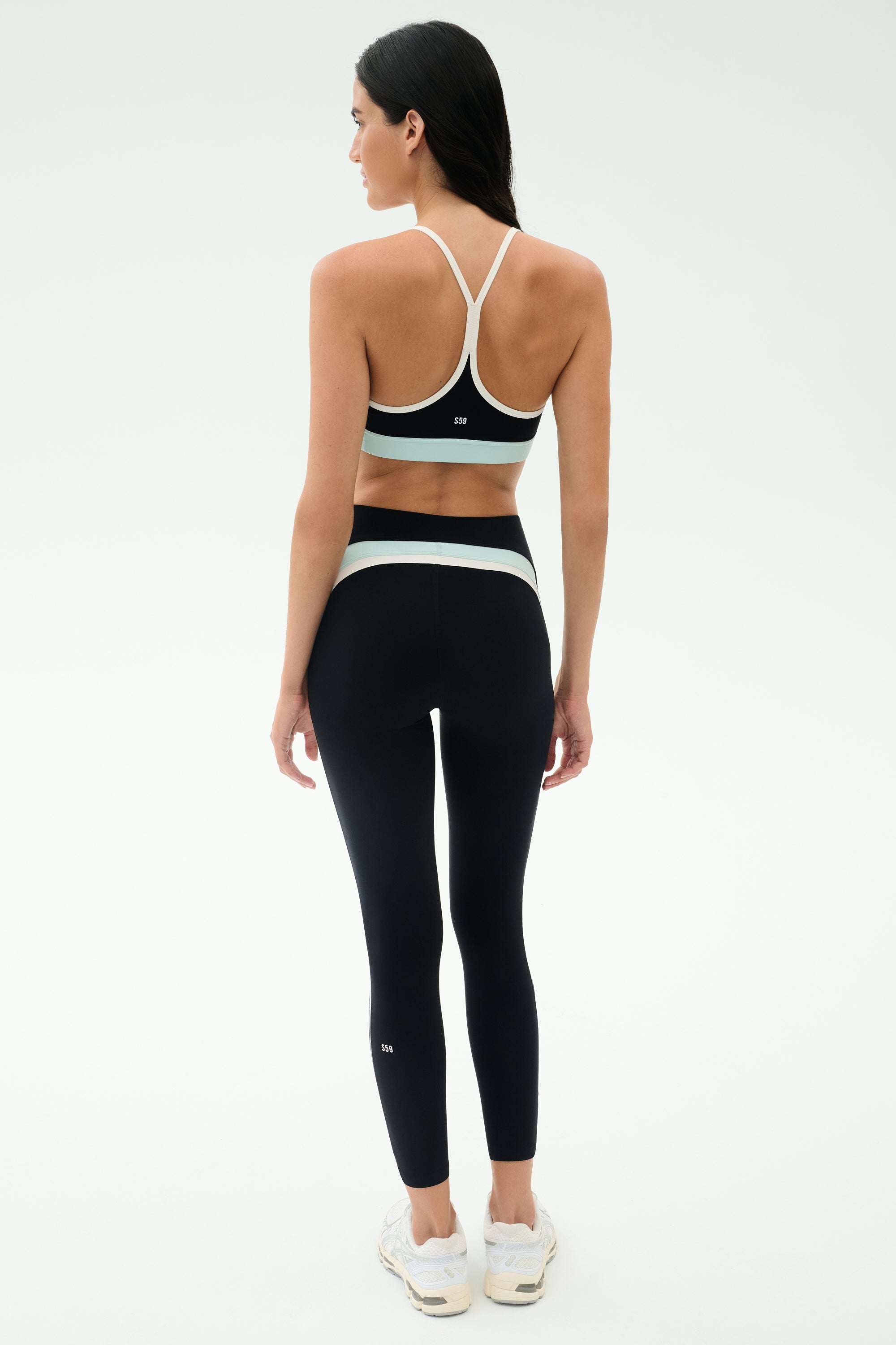 A woman faces away in a black SPLITS59 Emerson Airweight High Waist - Black/Ice legging with white accents and a black sports bra, paired with white sneakers—ideal for Pilates or hot yoga—against a plain white background.