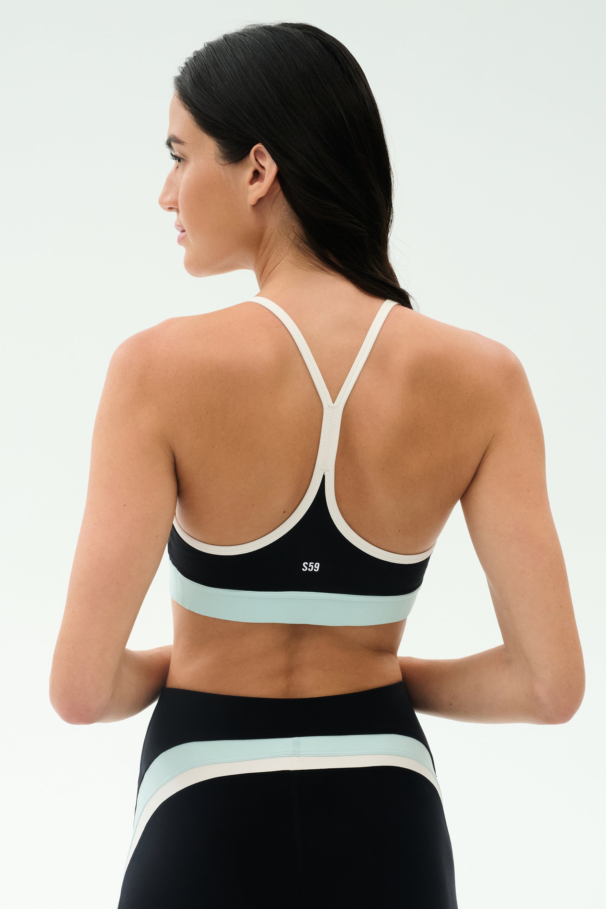A woman with long dark hair is shown from the back wearing the SPLITS59 Emerson Airweight Bra in Black/Ice with matching leggings—perfect for Pilates or hot yoga.