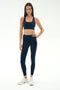 A woman stands against a plain background wearing the SPLITS59 Lexi Rigor Bra in Indigo/Blush, matching leggings with a side stripe, and white sneakers.