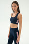 A woman wears the SPLITS59 Lexi Rigor Bra in Indigo/Blush, paired with matching high-waisted leggings featuring pink side stripes, posing against a plain light background.