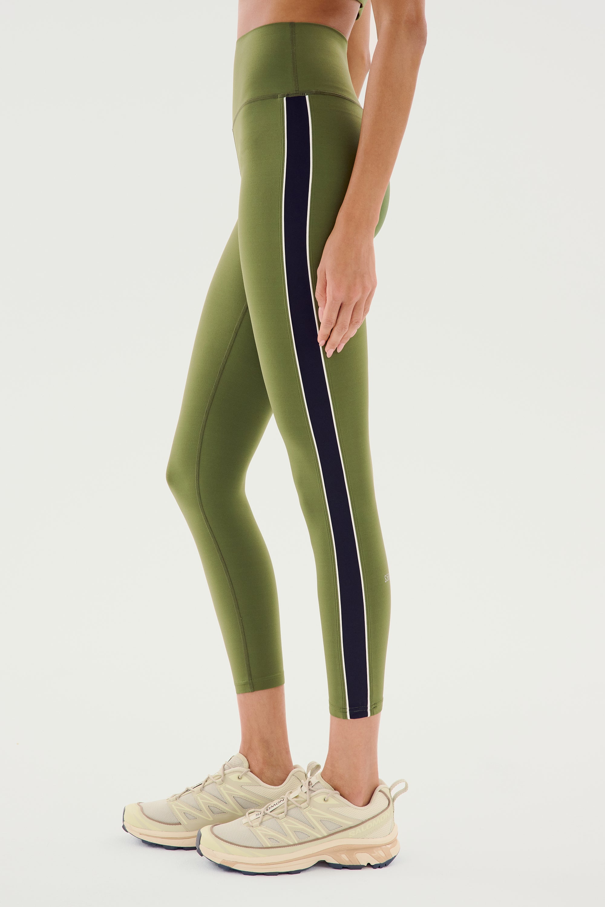 A person wearing the SPLITS59 Margot Rigor Legging in Olive/Black, made of spandex with a black vertical stripe on the side, and beige sneakers, stands against a plain background.