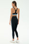 A woman stands facing away, wearing SPLITS59's Ella High Waist Airweight 7/8 leggings in Black/Stone Blue with white sneakers—ideal for hot yoga—against a plain white background.