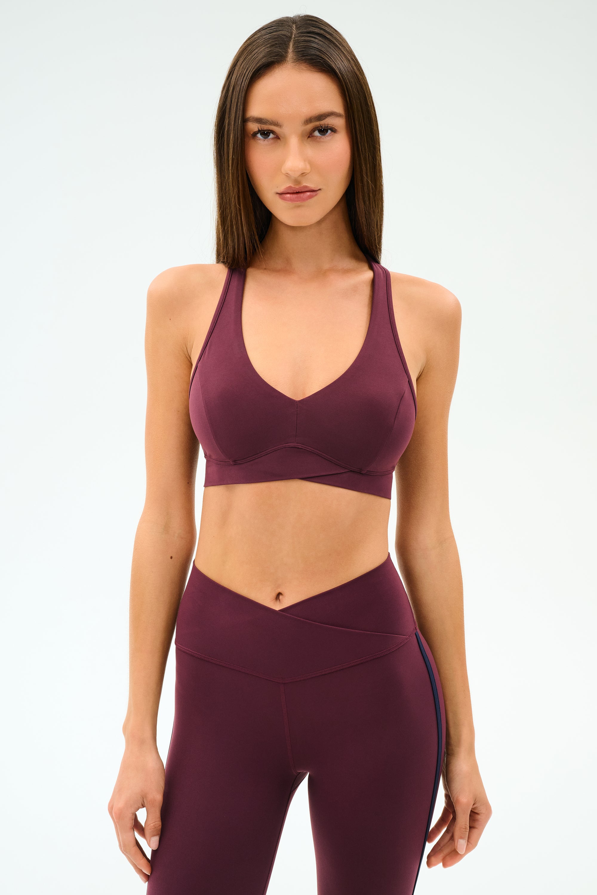 Woman stands against a plain background, wearing the SPLITS59 Mia Rigor Bra in Dark Cherry paired with matching high-waisted leggings.