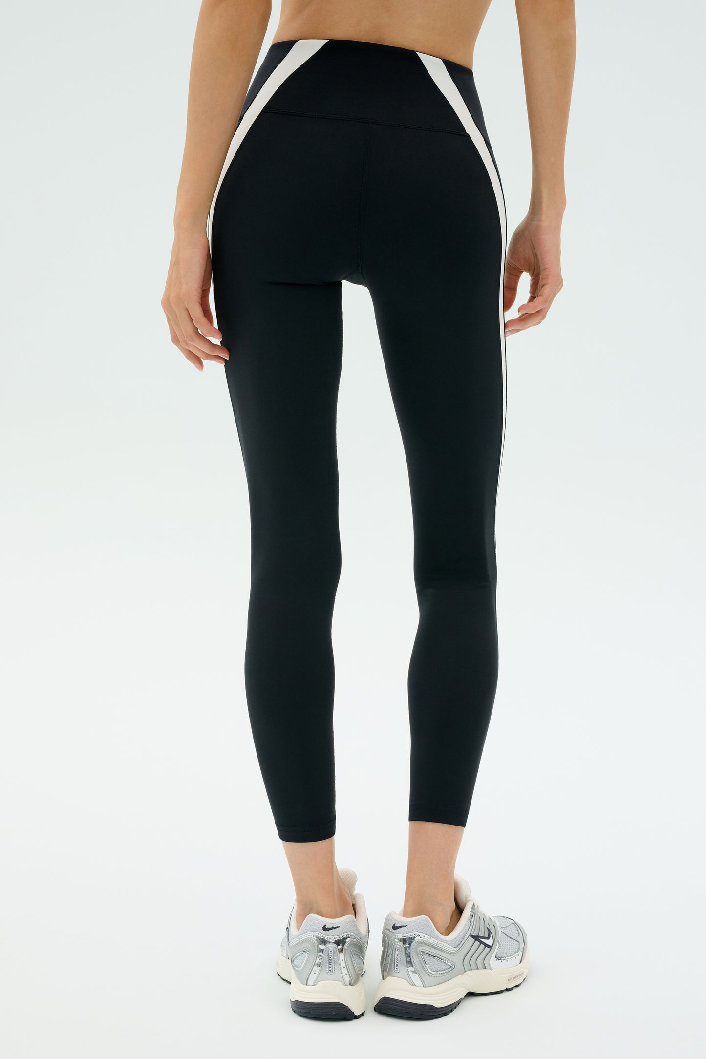 A person stands with their back to the camera, wearing SPLITS59 Miles High Waist Rigor 7/8 leggings in black/white, featuring white side stripes, paired with white athletic shoes.