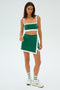 Wearing a green and white SPLITS59 Monah Rigor Bra - Arugula/White, along with an asymmetrical skirt, white sneakers, and socks, a person poses against a plain backdrop ready for their gym workout.