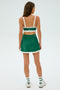 An individual with long hair stands with their back to the camera, wearing a SPLITS59 Monah Rigor Skort in Arugula/White and white sneakers, set against a plain backdrop.