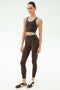 A woman models the SPLITS59 Amber Airweight Bralette in Dark Chocolate/Classic Blue, crafted from performance fabric perfect for hot yoga, with matching leggings and sneakers, posed against a white background.