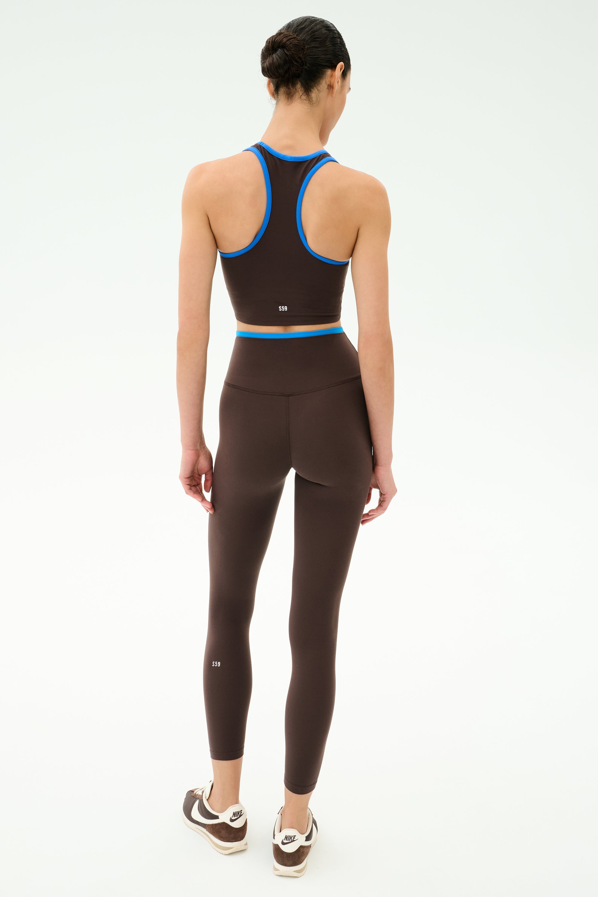 A woman faces away in the SPLITS59 Amber Airweight Bralette in Dark Chocolate/Classic Blue with matching leggings—ideal activewear for hot yoga—against a white background.