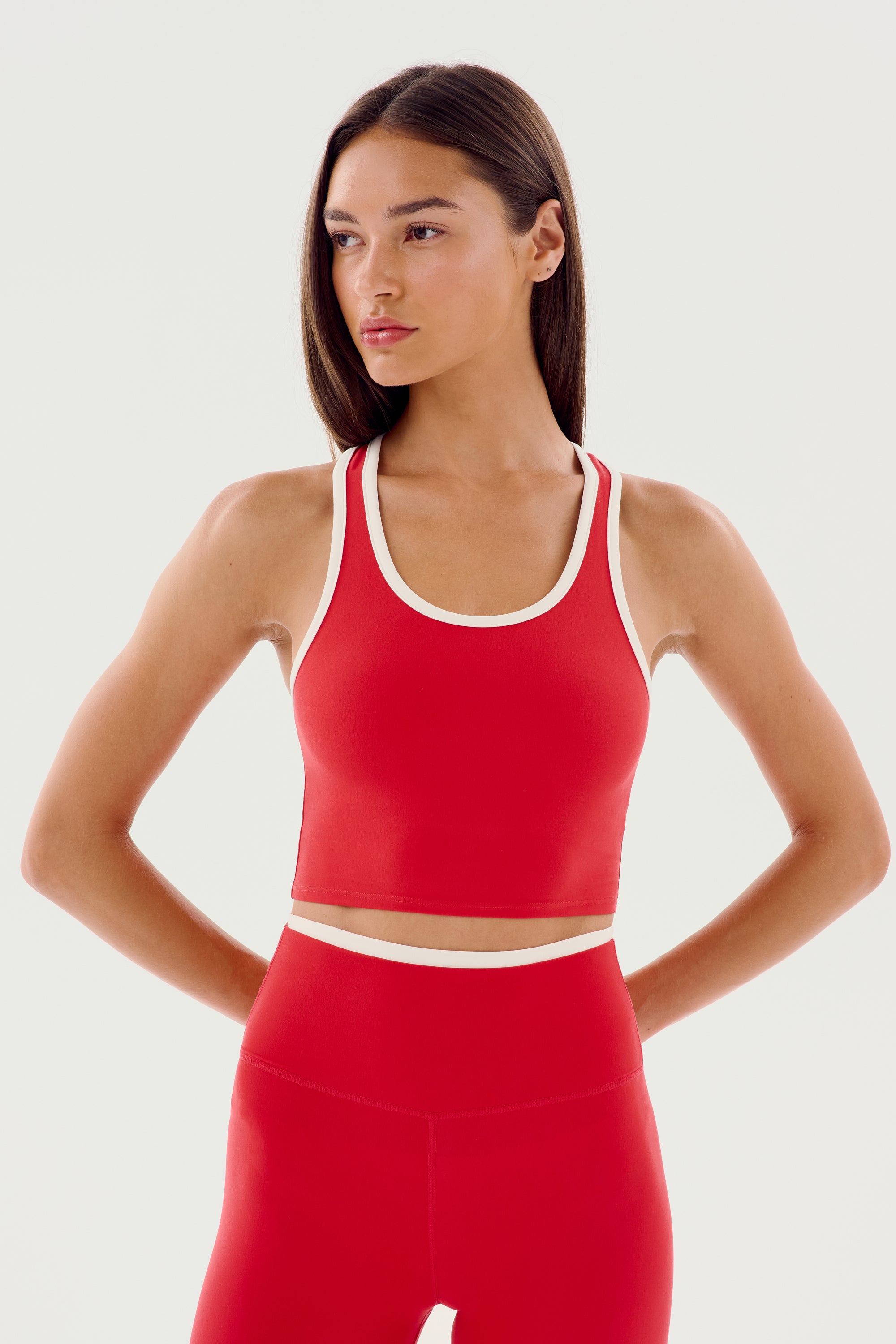 Person wearing a red athletic top and matching red bottoms with white trim, standing against a plain white background with hands on hips. The outfit is the SPLITS59 Amber Airweight Bralette - Pirate Red/White, perfect for hot yoga sessions.