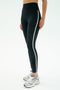 A person wears SPLITS59 Amber Airweight High Waist 7/8 leggings in Black/White, featuring a white side stripe, paired with white athletic sneakers and standing against a plain light background.