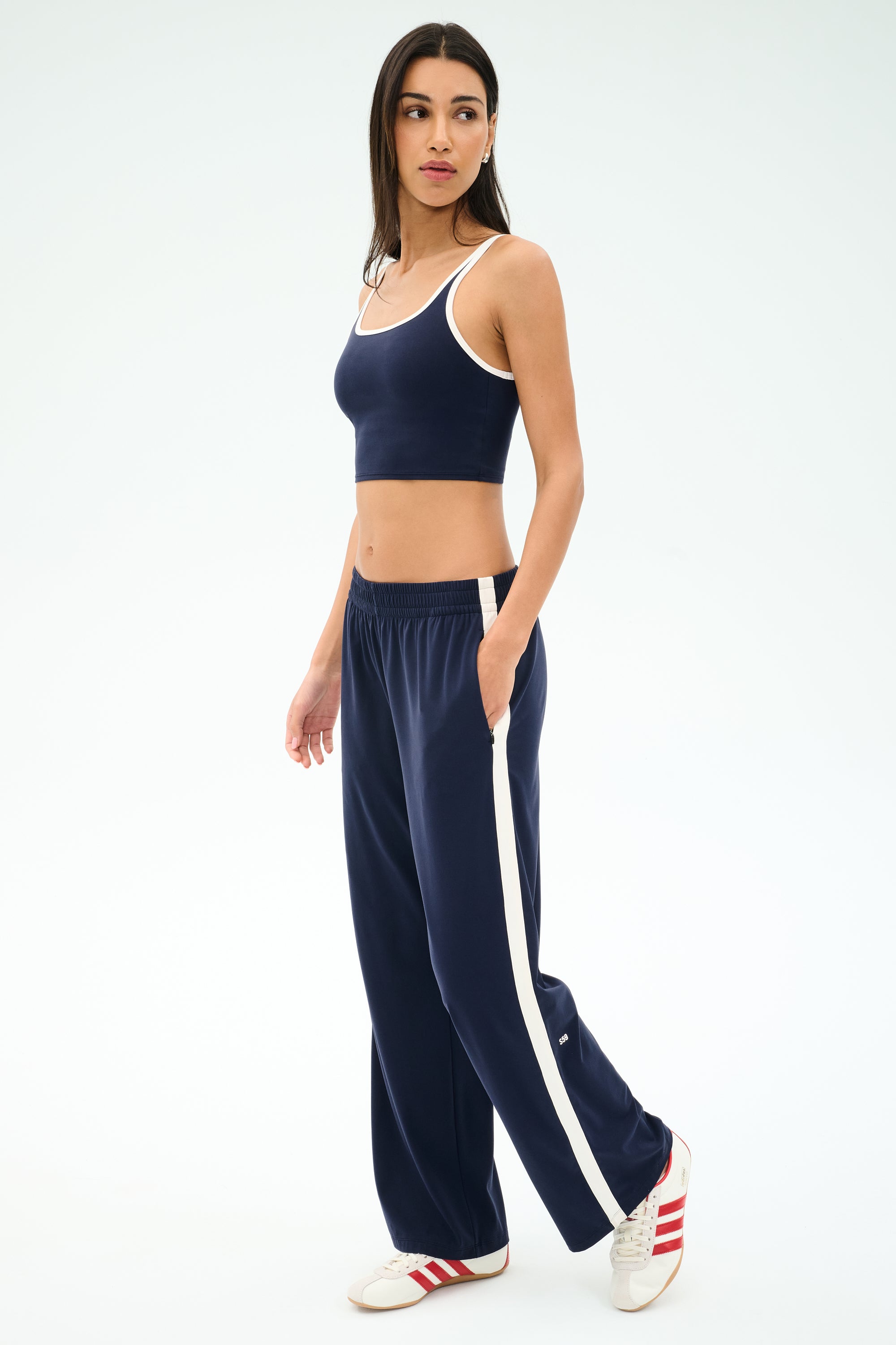 A woman in a navy crop top and SPLITS59 Maxie Rigor Track Pant - Indigo/White, wide-leg pants with white stripes, paired with red and white sneakers, stands against a plain background.