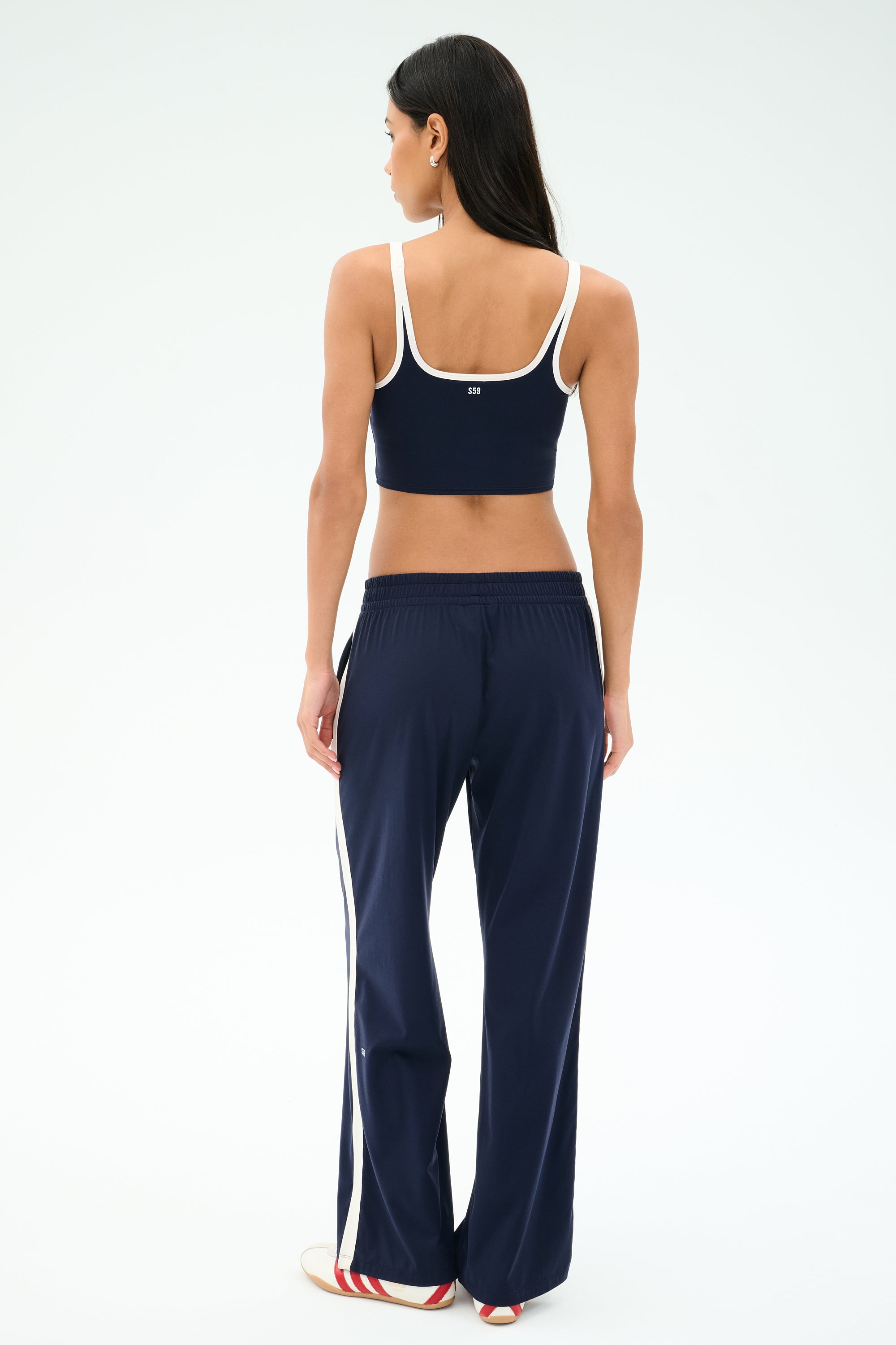 A woman, facing away, wears a navy blue crop top and SPLITS59 Maxie Rigor Track Pant in Indigo/White with white trim and side stripes, against a plain white background.