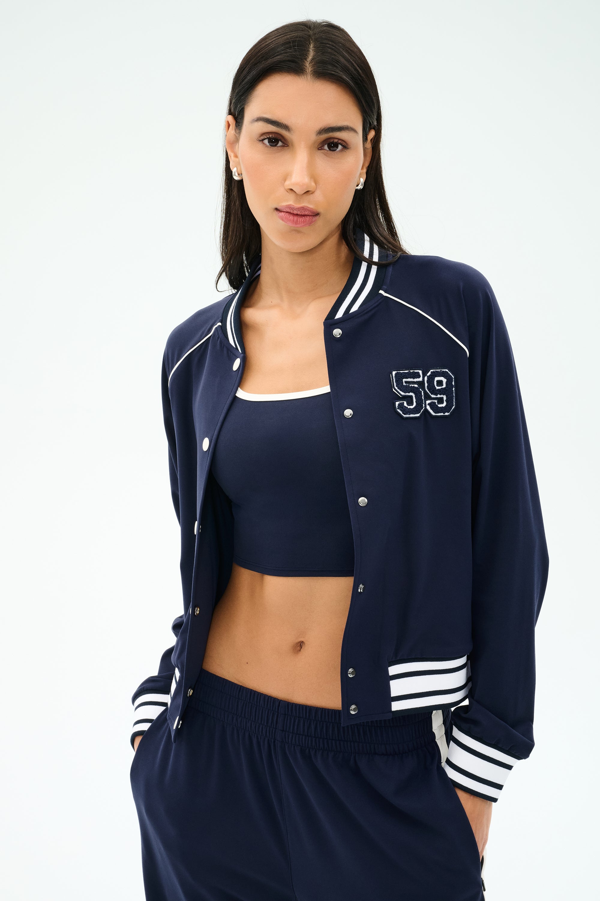 A woman stands against a plain light background wearing the SPLITS59 Hayes Varsity Rigor Jacket in Indigo, paired with a matching stretch crop top and pants.