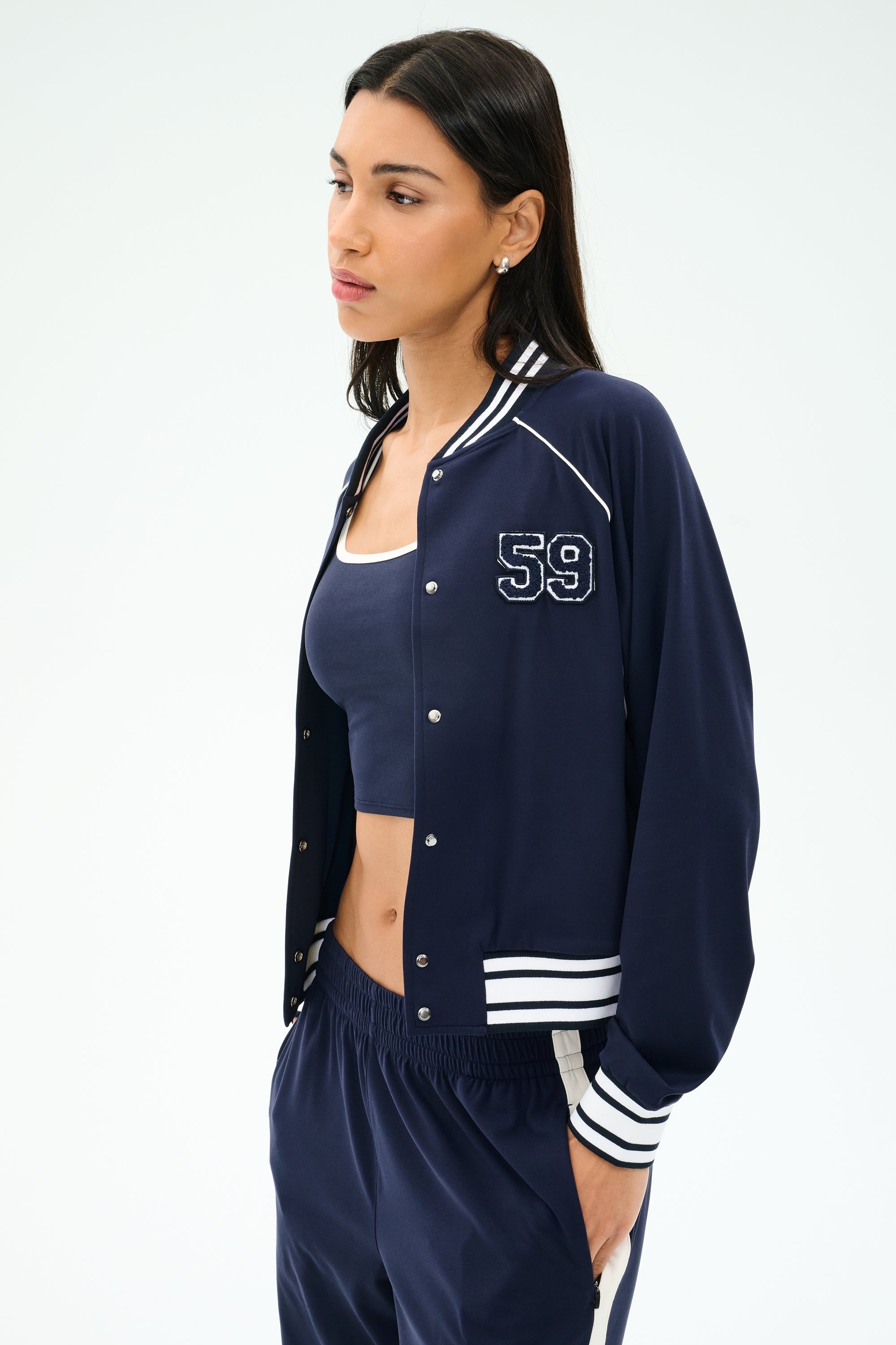 A woman stands against a plain light background, wearing the SPLITS59 Hayes Varsity Rigor Jacket in indigo with “59” on the chest, a matching crop top, and navy stretch track pants.
