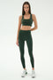 A woman stands against a plain background wearing the SPLITS59 Margot Rigor Legging in Military/Blush and a matching dark green sports bra, paired with white sneakers.