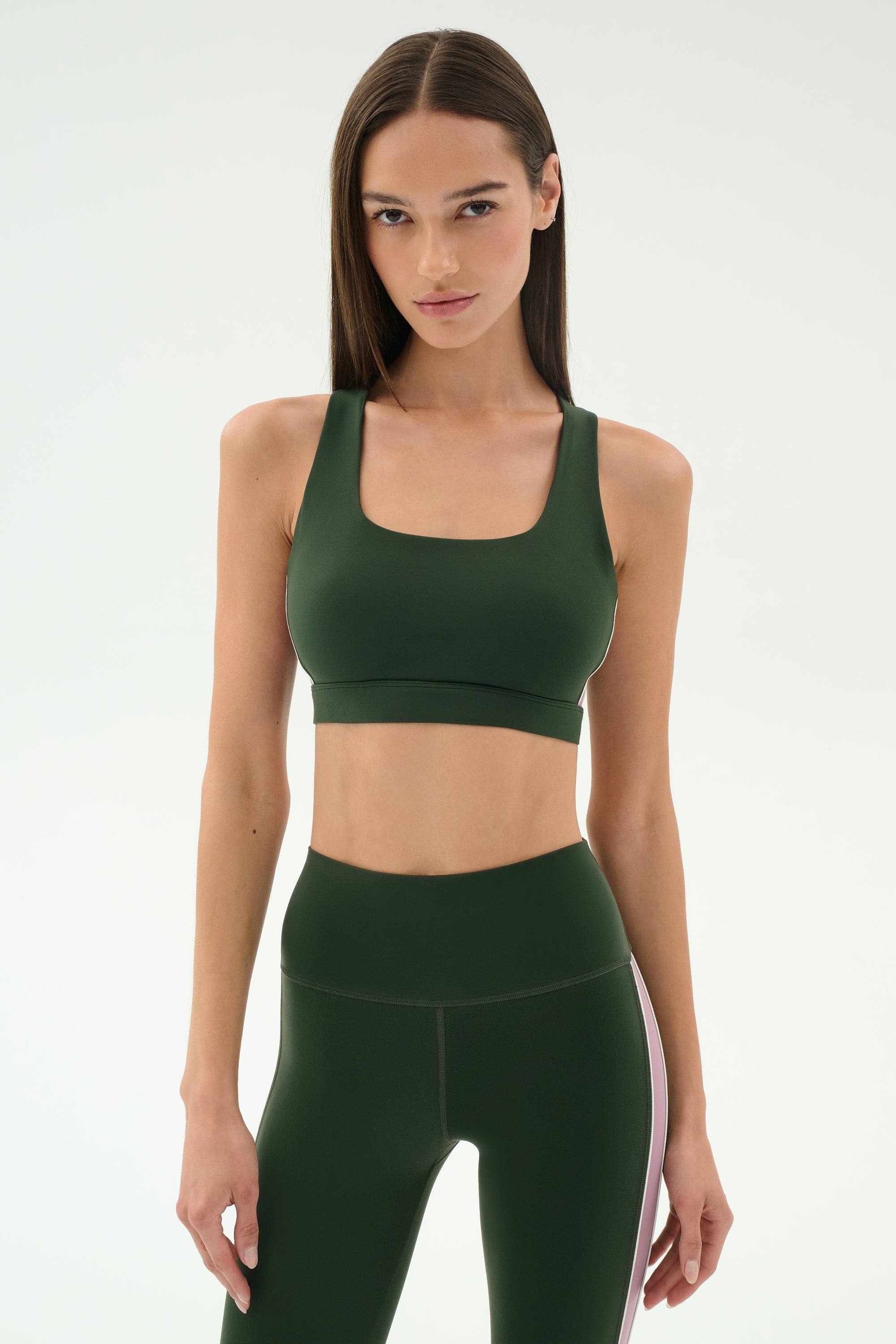 A woman faces forward, wearing the SPLITS59 Lexi Rigor Bra in Military/Blush and matching high-waisted leggings with a white side stripe, set against a plain light background.
