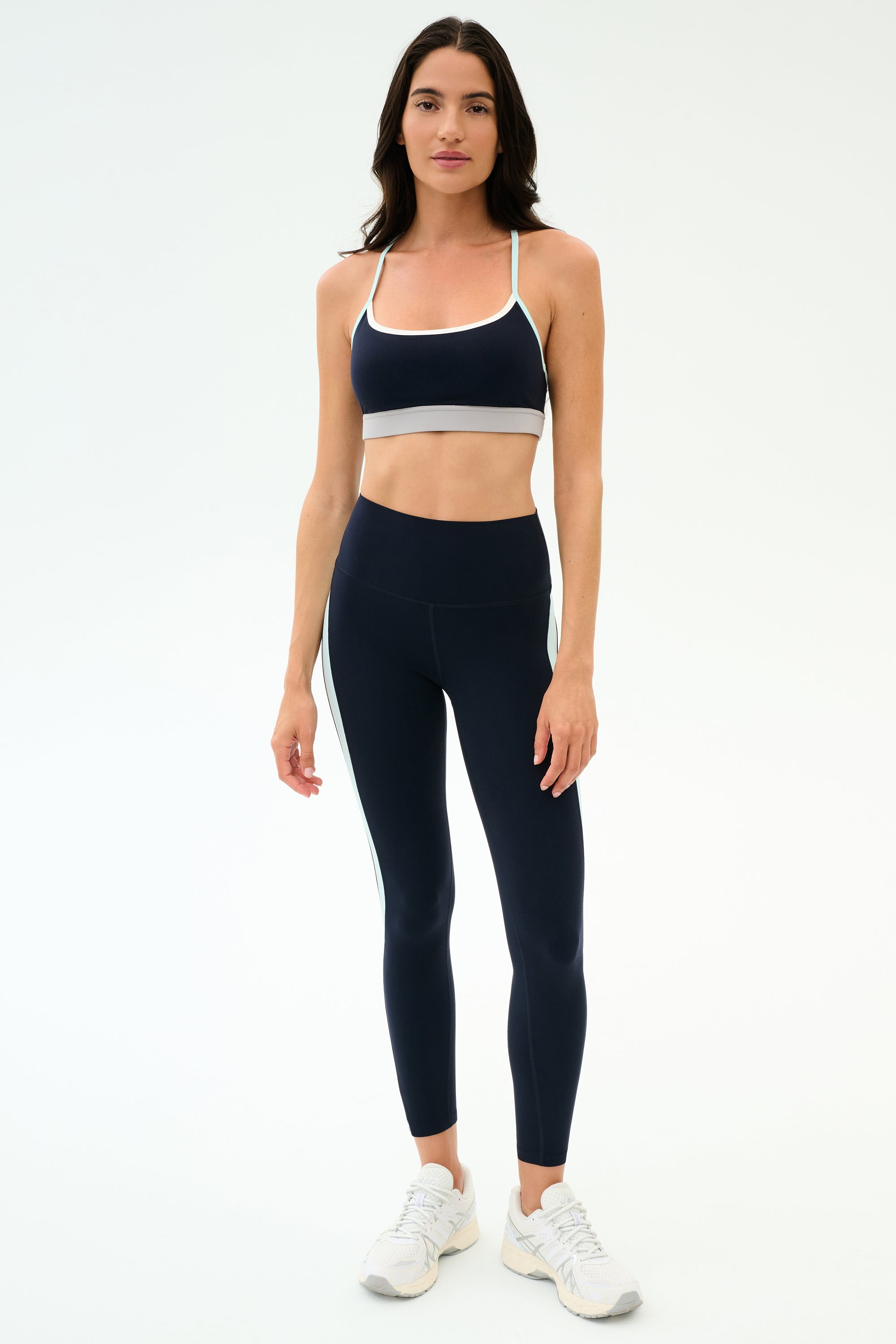 A woman stands facing forward in SPLITS59's Emerson Airweight High Waist leggings in Indigo/Dove Grey with a matching sports bra and white sneakers—perfect for Pilates or hot yoga—against a plain light background.