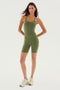 A woman with long hair wearing a body-hugging Olive Airweight 6” Short Jumpsuit by SPLITS59 and white sneakers stands against a plain white background.