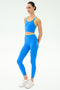 A woman stands with hands behind her back, wearing SPLITS59's Dual High Waist Airweight 7/8 leggings in Classic Blue/Ray with a matching crop top and white sneakers, against a plain white background.