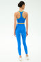 A woman stands facing away in SPLITS59's Dual High Waist Airweight 7/8 leggings and matching sports bra in Classic Blue/Ray with yellow trim, made from ultra luxe fabric, paired with white sneakers against a white background.