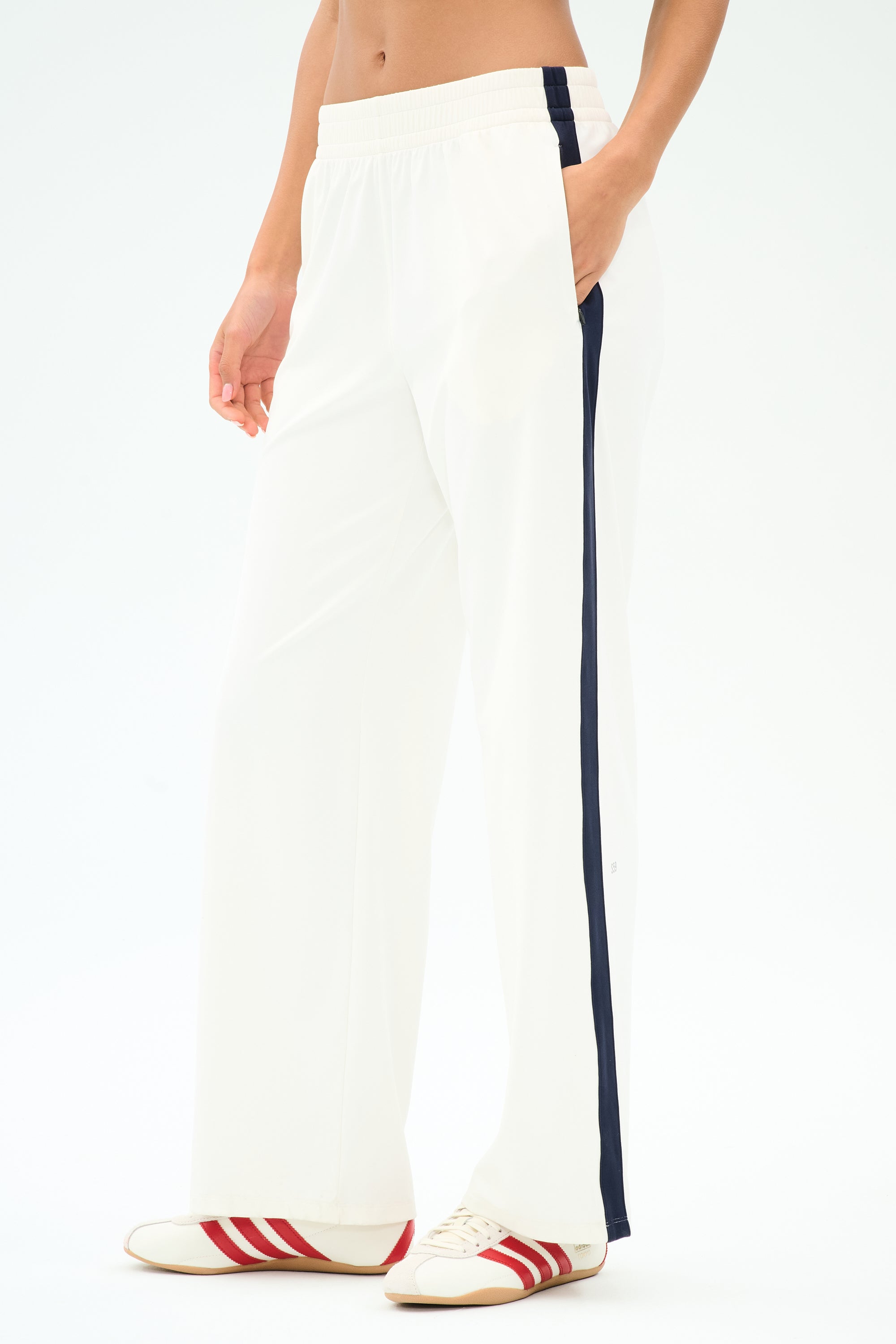 A person wears SPLITS59 Maxie Rigor Track Pant in White/Indigo, featuring a navy side stripe, paired with white sneakers with red stripes, standing against a plain background.