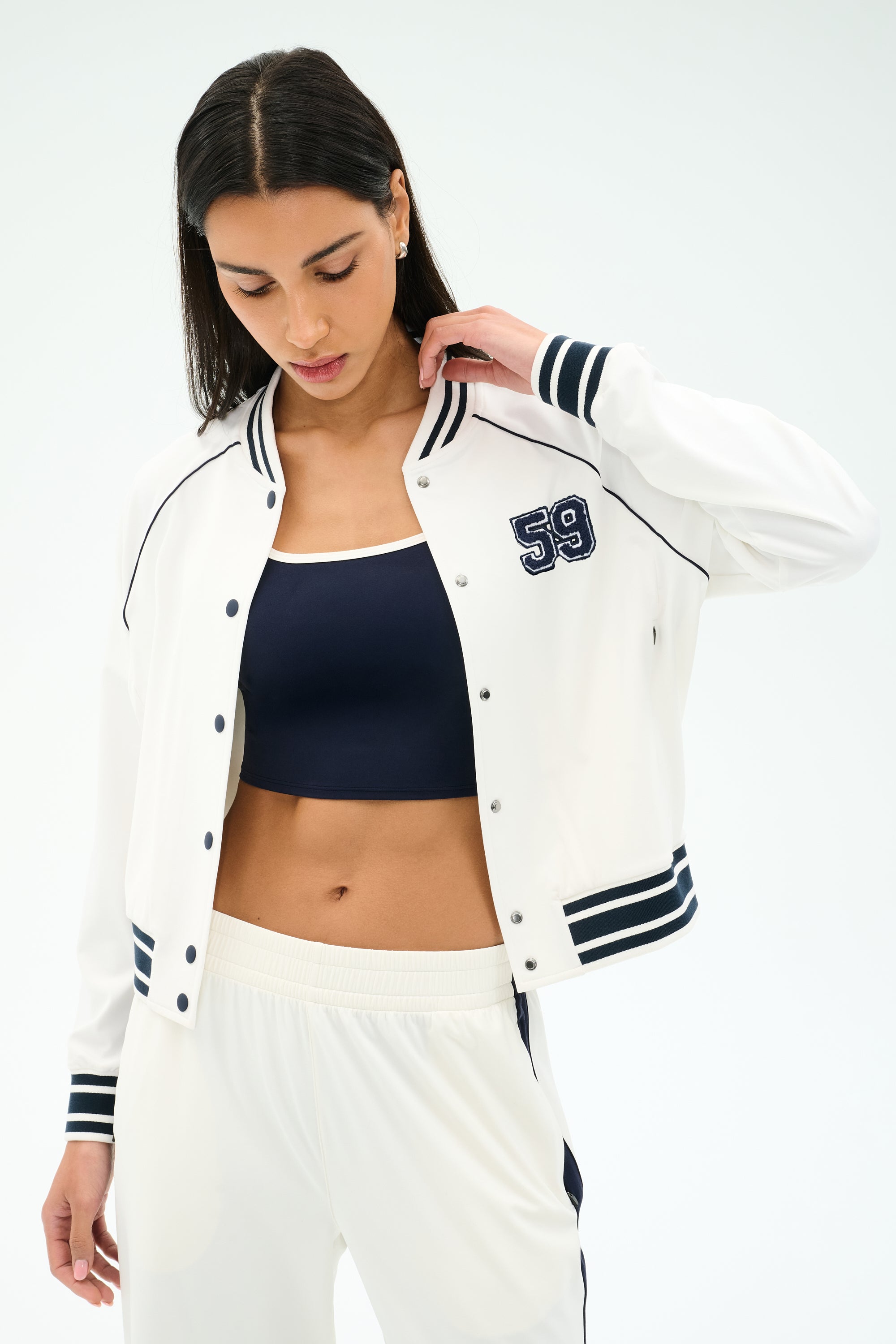 A woman wearing the SPLITS59 Hayes Varsity Rigor Jacket in white, styled over a navy sports bra and white track pants, looks down while touching her hair.