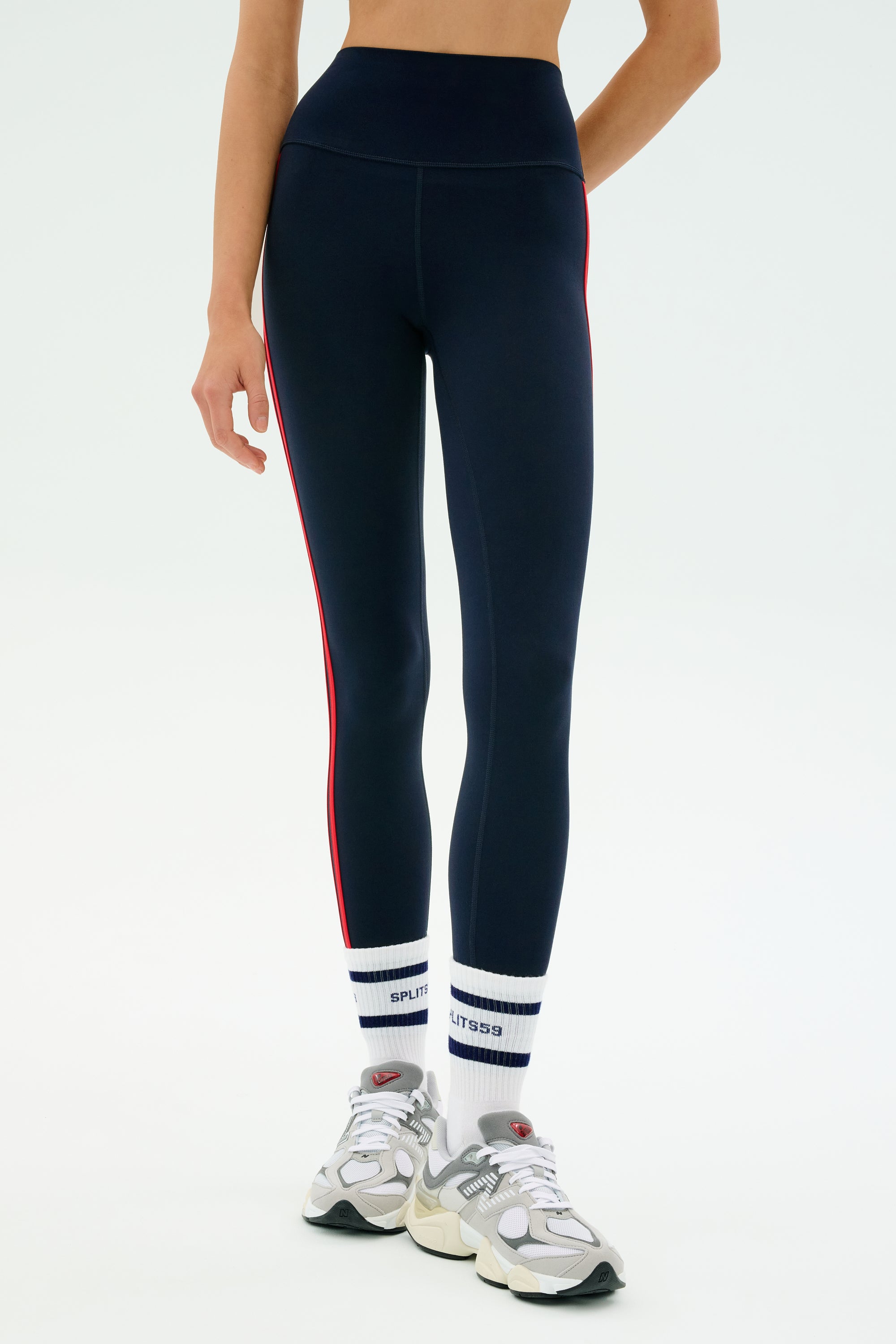 A person wearing SPLITS59 Ella High Waist Airweight 7/8 leggings in Indigo/Pirate Red, paired with white and navy socks and white sneakers—perfect for hot yoga—stands against a plain background.