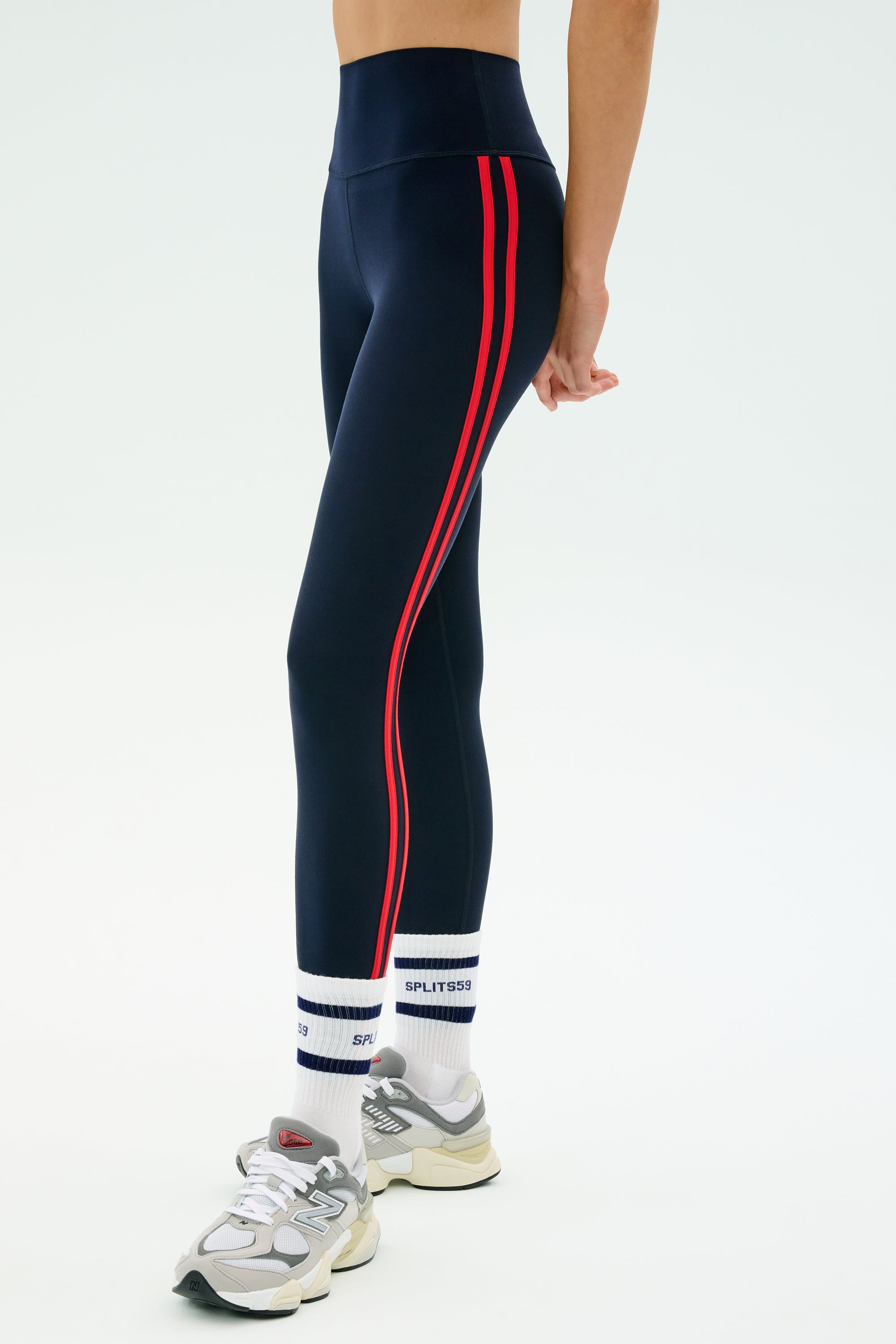 A person wearing SPLITS59 Ella High Waist Airweight 7/8 leggings in Indigo/Pirate Red, paired with white socks with blue text and gray sneakers, stands against a plain light background.
