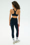 A woman with curly hair, seen from behind, wears the SPLITS59 Ella High Waist Airweight 7/8 leggings in Indigo/Pirate Red, paired with a black sports bra, white socks, and sneakers against a plain light background.