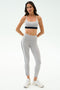 A woman poses against a plain background in the SPLITS59 Emerson Airweight Bra in Dove Grey/Indigo, paired with matching leggings and white sneakers—perfect for Pilates or hot yoga.
