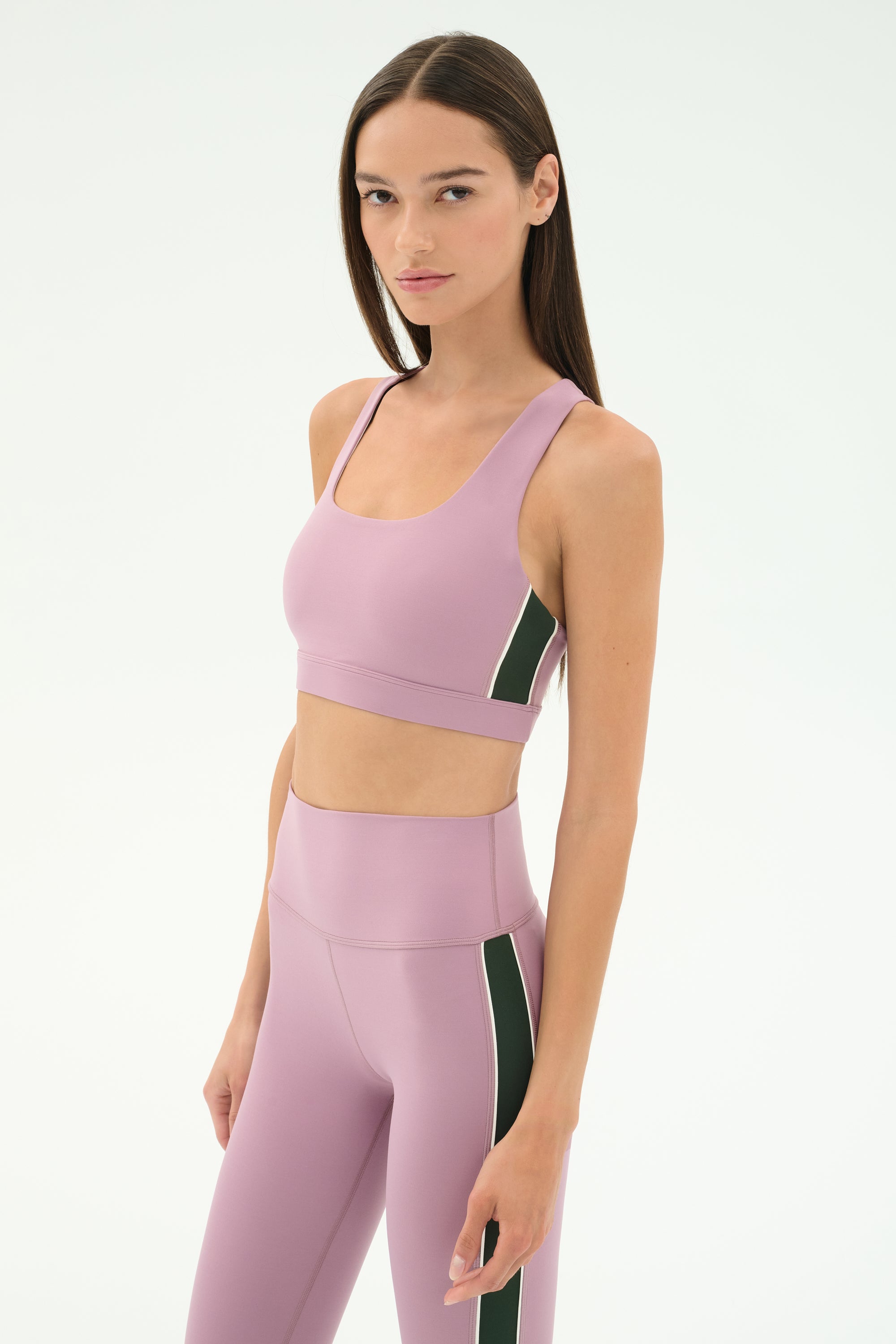 A woman wears the SPLITS59 Lexi Rigor Bra in Blush/Military with matching high-waisted leggings featuring green and white side stripes, standing against a plain white background.