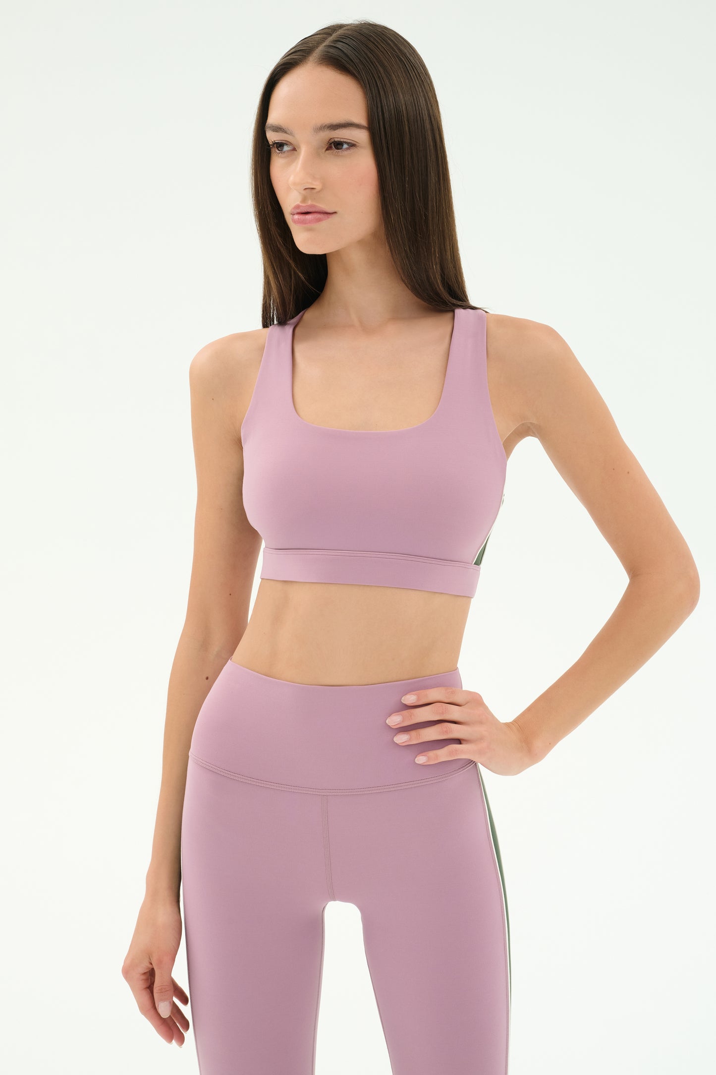 A woman wearing the SPLITS59 Lexi Rigor Bra in Blush/Military and matching high-waisted leggings stands against a plain white background.