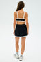 A person, back to camera, wears a black and white SPLITS59 Monah Rigor Bra with matching skirt and sneakers. This sleek outfit is ideal for gym workouts against a plain white backdrop.