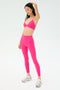 A person stands in a studio wearing SPLITS59's Sprint High Waist Rigor 7/8 leggings in Berry, paired with a matching pink sports bra and white athletic shoes against a plain light background.