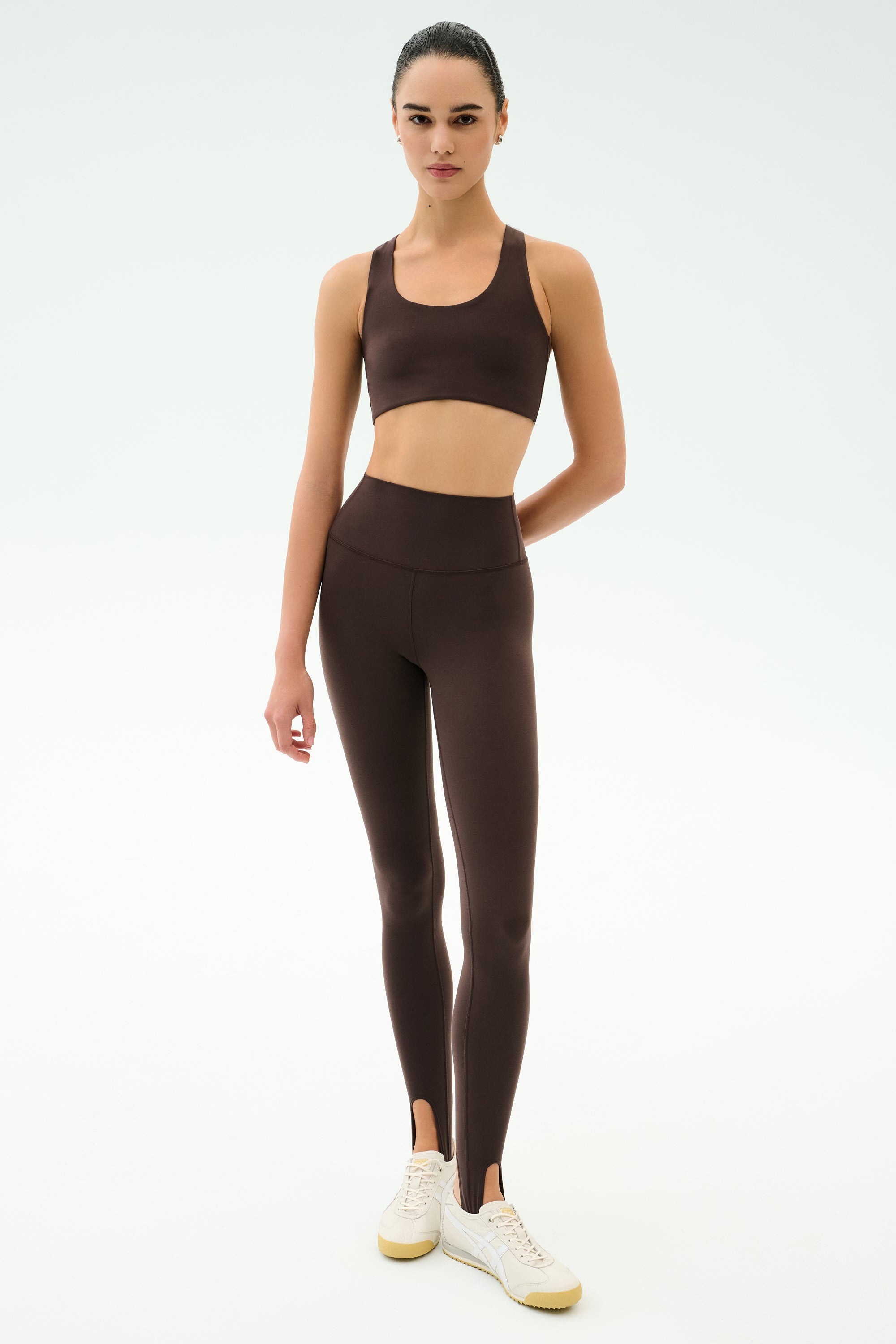 A person faces forward wearing the SPLITS59 Sara Airweight Bra in Dark Chocolate with matching high-waisted stirrup leggings and white sneakers, ready for Pilates or hot yoga, against a plain light background.
