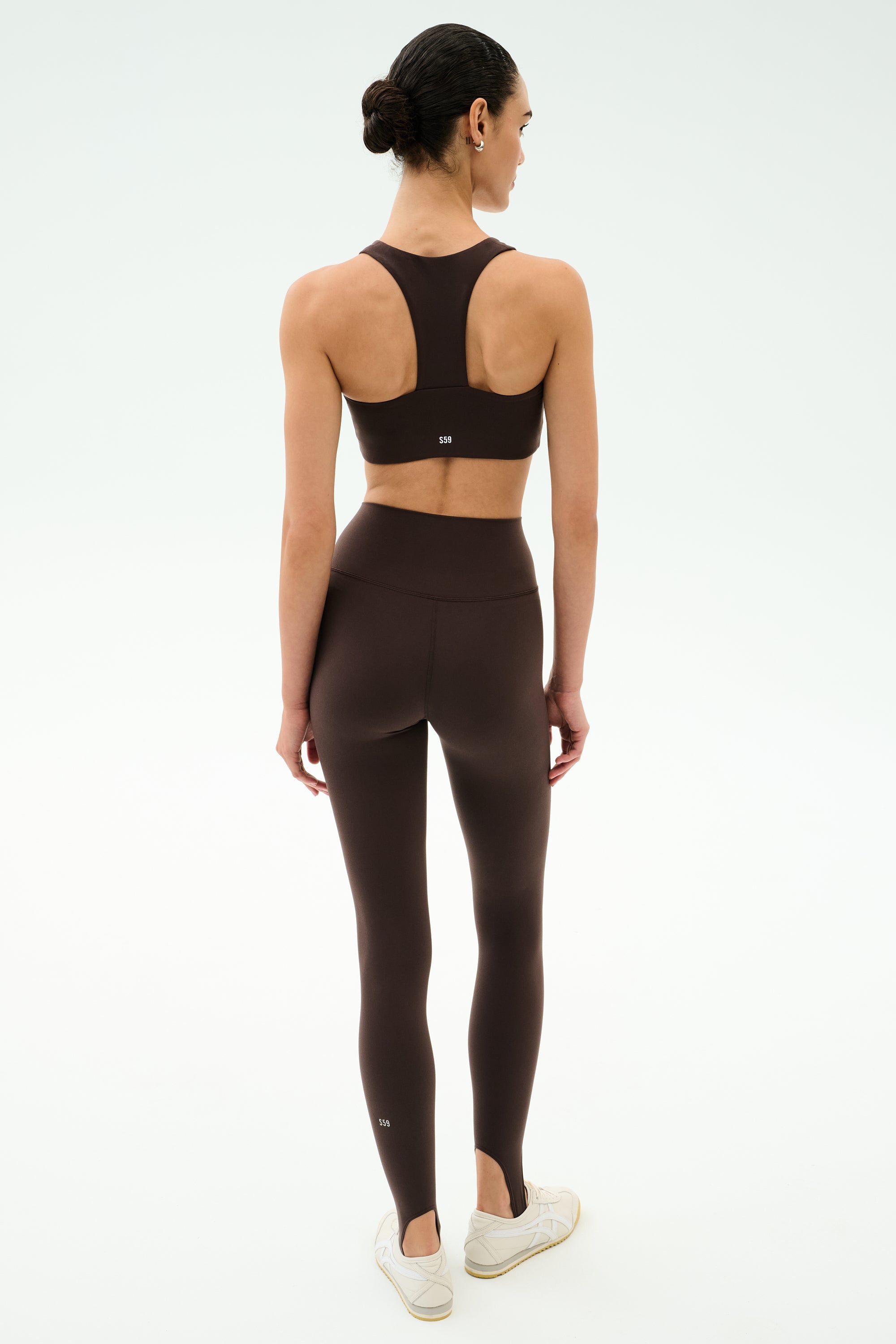 A woman stands facing away, wearing the SPLITS59 Sara Airweight Bra in Dark Chocolate with matching high-waisted leggings and white sneakers, perfect for Pilates, against a plain white background.