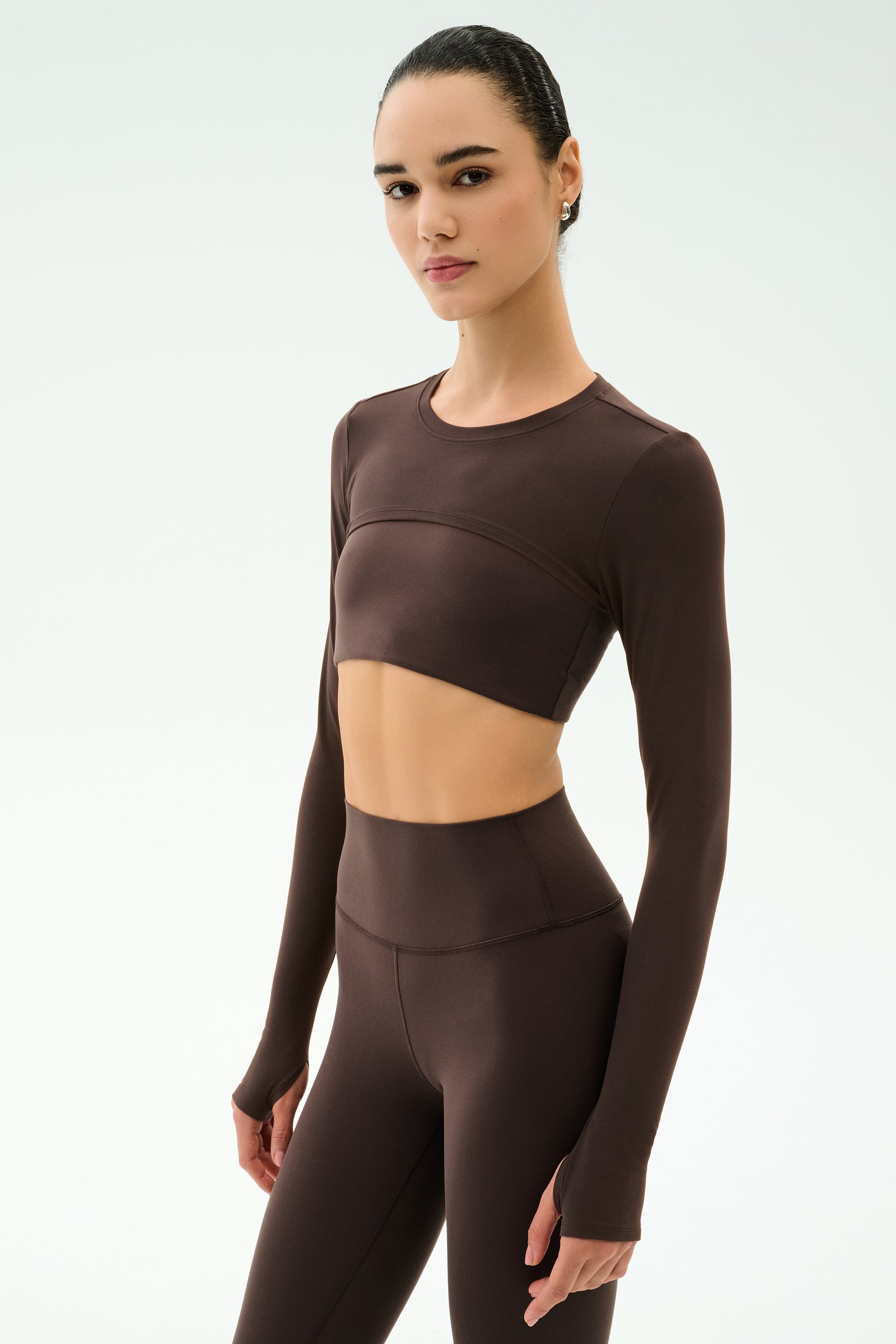 A woman stands against a plain light background in a matching dark brown long-sleeve crop top and high-waisted leggings, her yoga-ready look finished with the SPLITS59 Amelia Airweight Lite Shrug in Dark Chocolate.