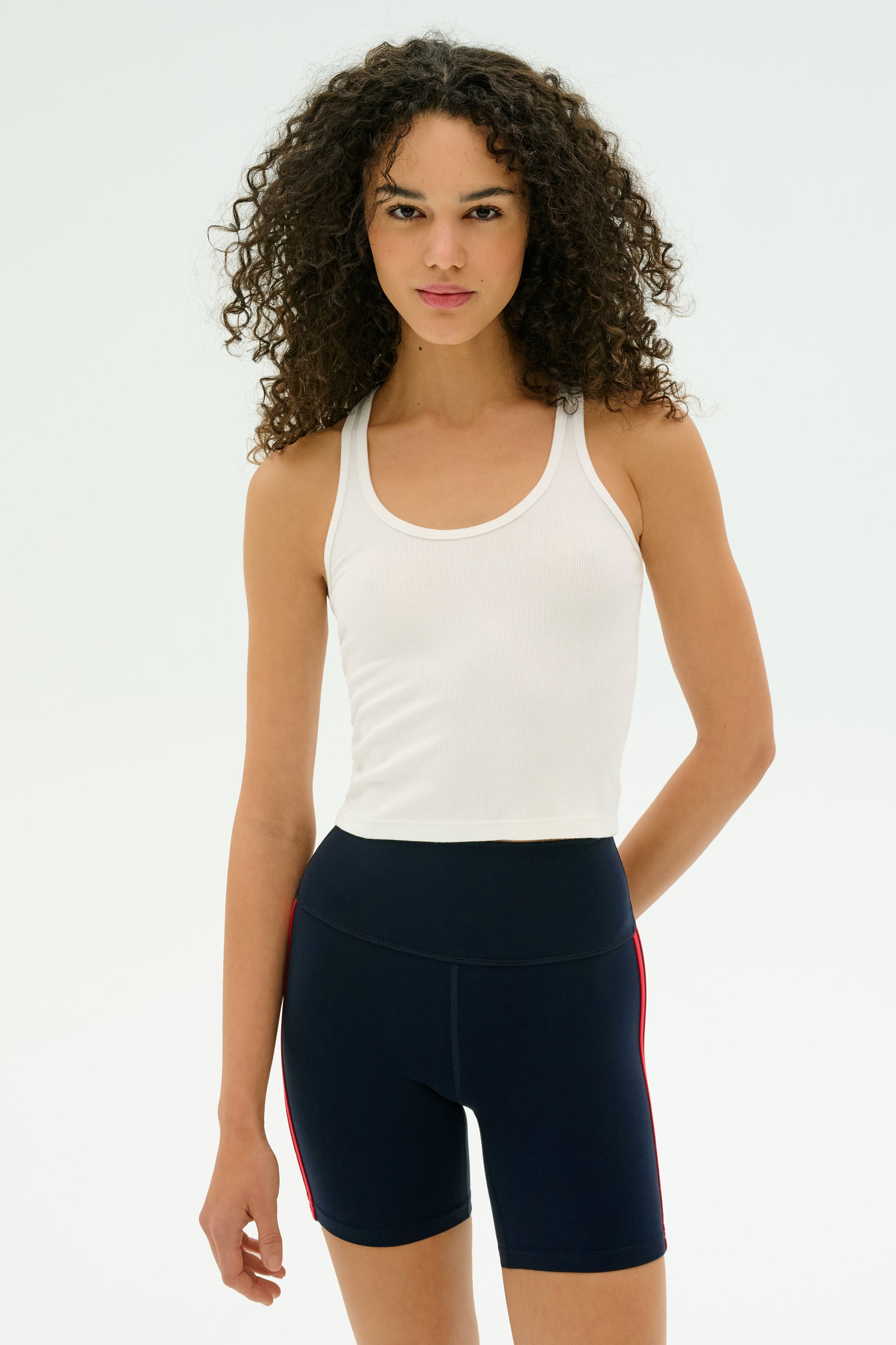 A person with curly hair models the SPLITS59 Ashby Rib Crop - White, paired with navy bike shorts featuring red stripes, against a plain light background—perfect for gym workouts.